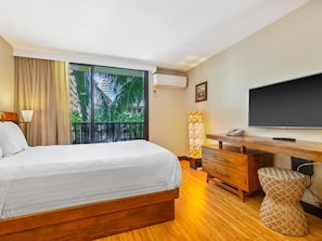 Hotelkamer met een bed, bureau, stoel, televisie en uitzicht op palmbomen door een groot raam.
