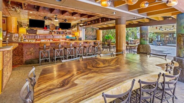 An outdoor bar area with a long wooden table and multiple chairs, surrounded by a rustic wooden ceiling and exposed beams.