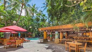 An outdoor seating area with wooden tables and chairs, a red umbrella, and a pool in the background.