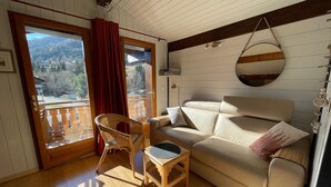 A cozy living room with a wooden floor, a beige sofa, a wicker chair, and a small table. A large window with a view of a forest and mountains is visible. - Attic Apartment 3 Rooms 74m², South Balcony, Private Parking, Wifi (La Clusaz)