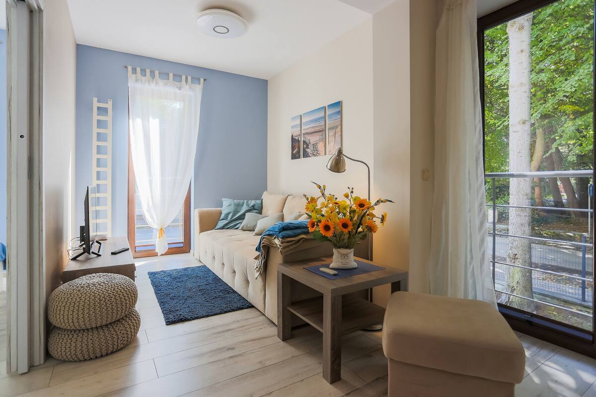 Een moderne woonkamer met een bank, een salontafel met een bloevas, een ladder en een groot raam met uitzicht op bomen.
