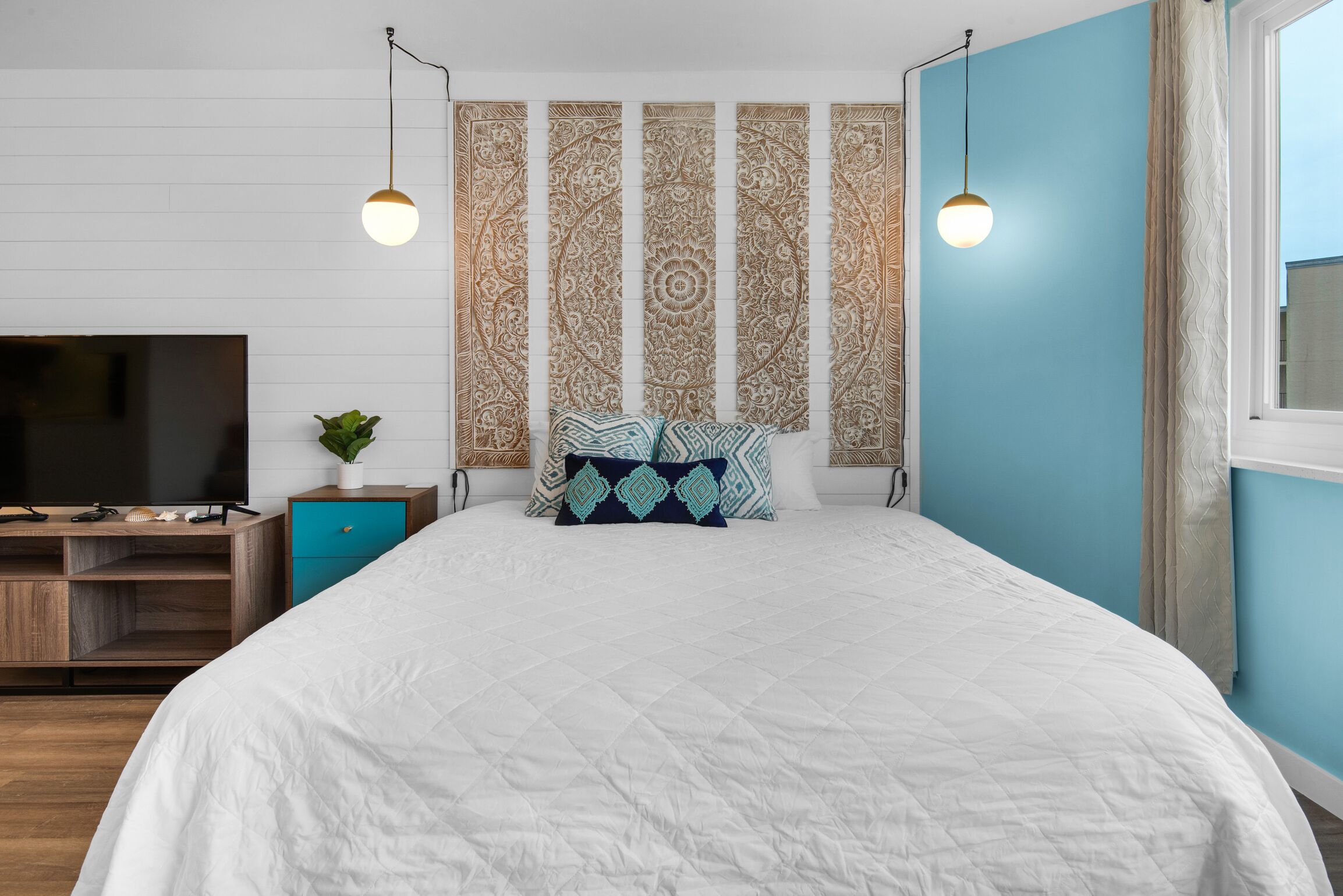 Ein Schlafzimmer mit einem großen Bett, einem Fernseher auf einem Holzboden, einer Pflanze und Wanddekoration mit Muster.