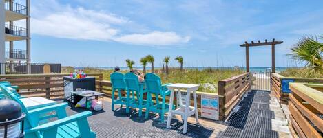 A beachside patio with outdoor seating, a wooden deck, and a view of the ocean.