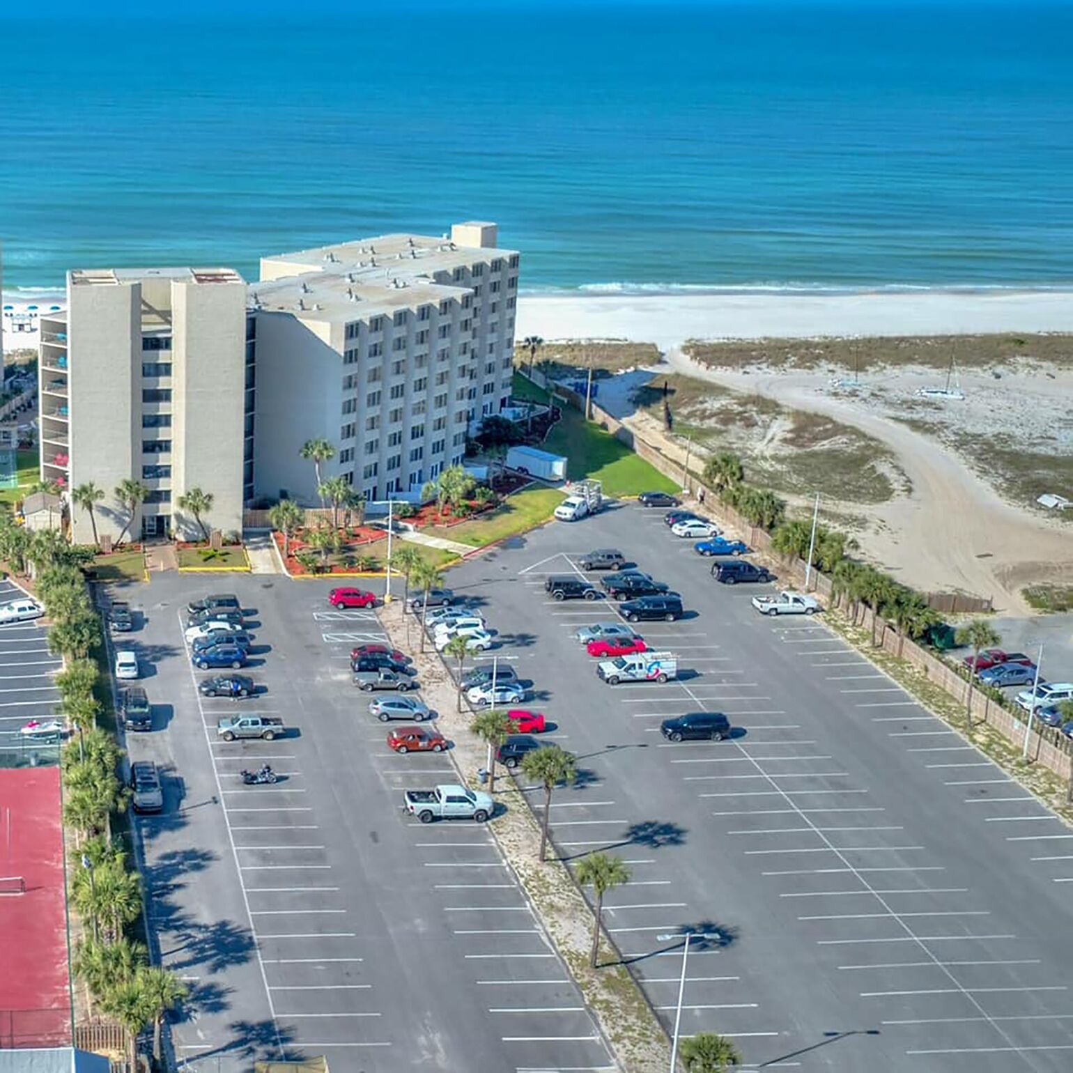A multi-story hotel with a parking lot in front, adjacent to a beach.