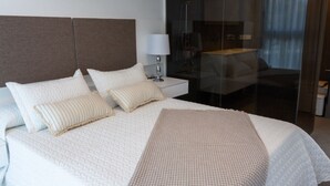 Ein Hotelzimmer mit einem großen Bett, zwei Nachttischen, einer Lampe und einer Glastrennwand, die den Raum von einem Essbereich abteilt.