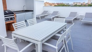 Eine Dachterrasse mit weißem Tisch und Stühlen, einem Grill und Blick auf die Stadt.