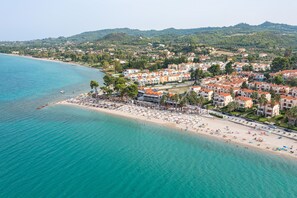 An aerial view of a coastal town with a beach, buildings, and a hilly landscape in the background. - Tridente Mare (Kassandra)