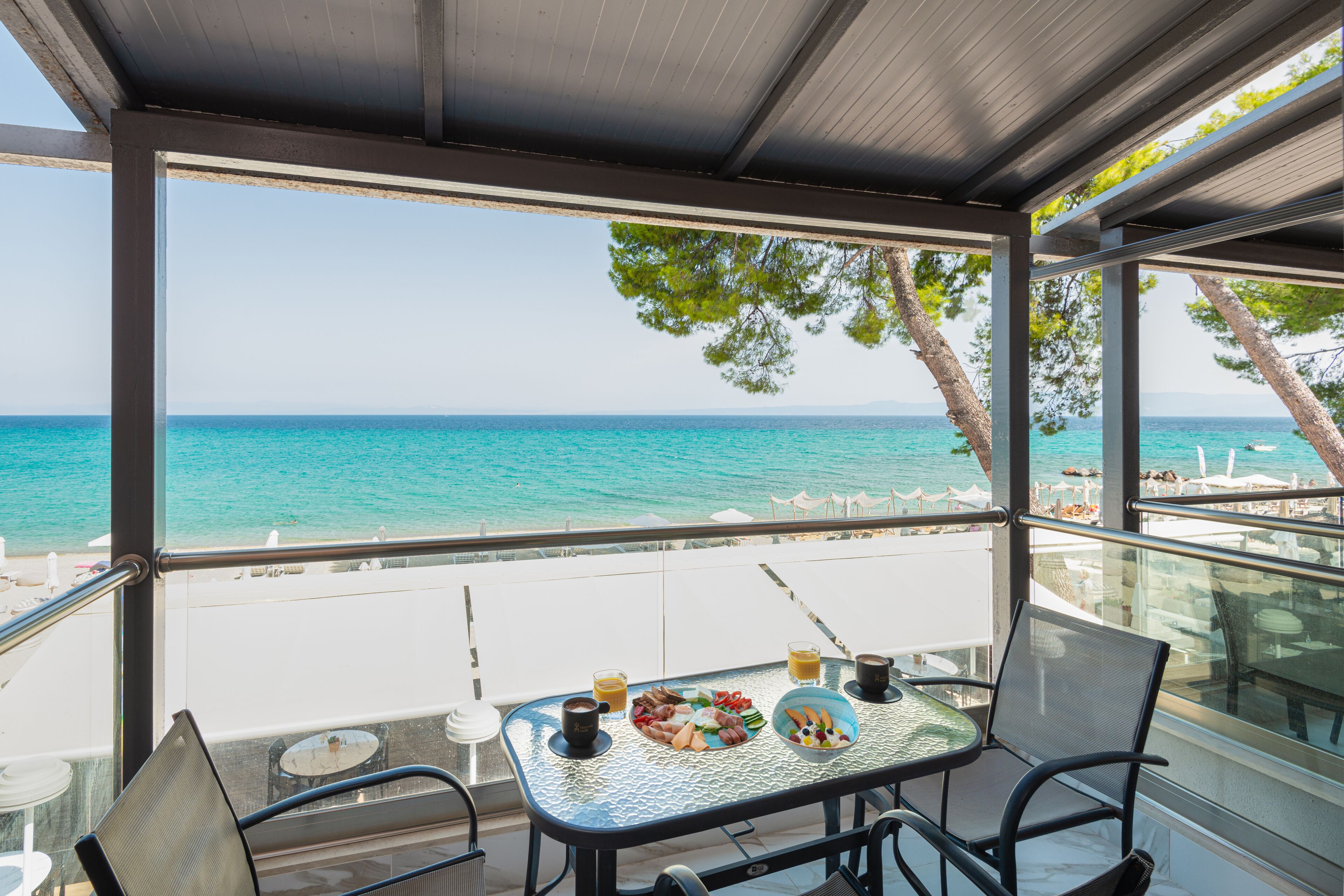 A balcony with a table set for breakfast, overlooking a beach with clear turquoise water and a white sandy shore.