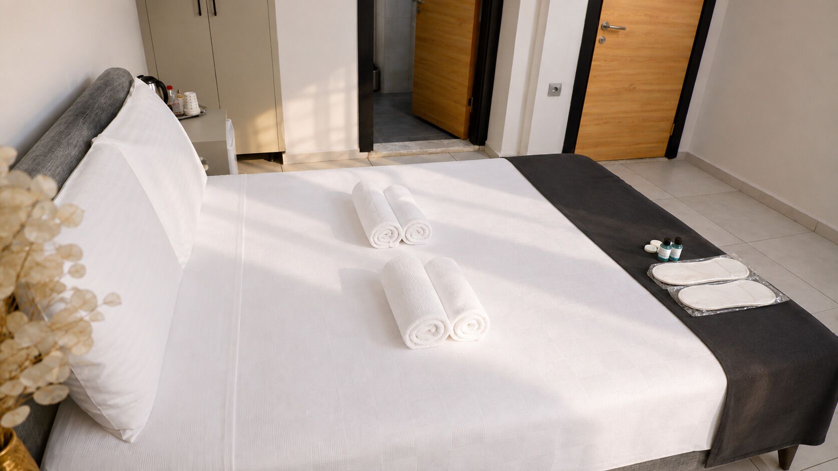 A neatly made bed with white linens, a dark-colored bedspread, and two rolled towels.