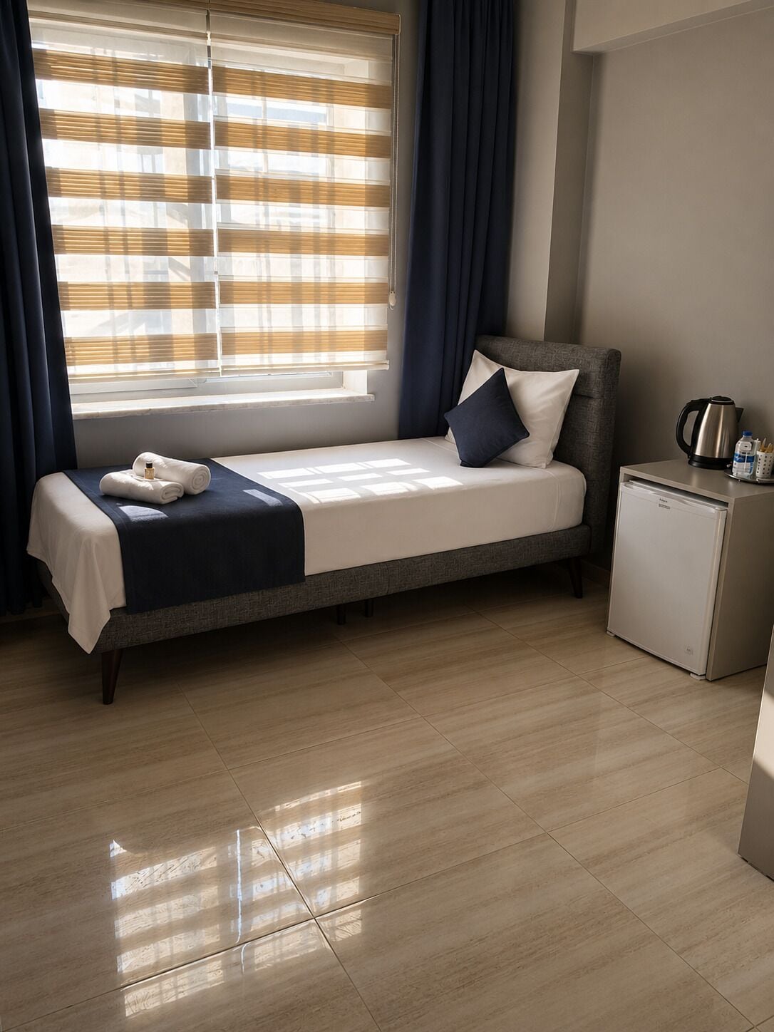 A single bed with a headboard, a bedside table with a kettle, and a small refrigerator.