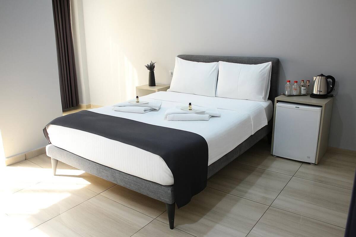 A modern hotel room with a bed, bedside tables, a small refrigerator, and a window with curtains.