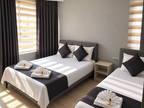 A hotel room with two beds, each with a grey headboard, white bedding, and black and white pillows. There are two bedside tables with lamps, and the room has a large window with blinds. - Caliskanlar Otel (Çanakkale)
