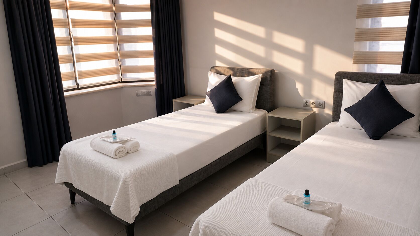 A hotel room with two single beds, a nightstand, a bedside table, and a window with blinds.
