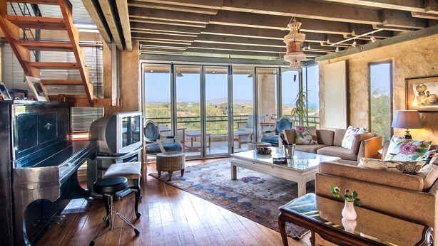 Ein geräumiges Wohnzimmer mit Holzboden, einem Fernseher, einer Couch und einer großen Schiebeglastür, die auf einen Balkon mit Blick ins Grüne führt.