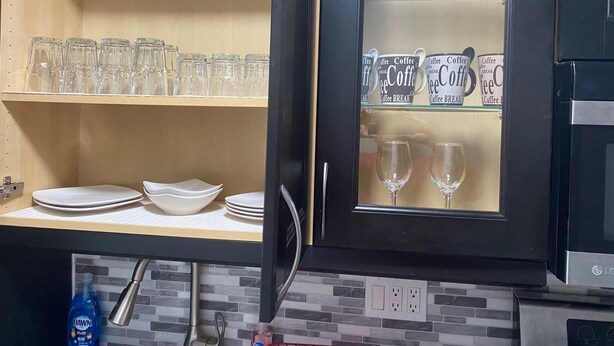 Een keuken met een zwart kastje met een glazen vitrine voor koffiemokken en glazen. Daarboven staan planken met heldere glazen en borden.