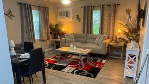 Een moderne woonkamer met een grijze bank, een salontafel en een eethoek met een zwarte tafel en stoelen. Er zijn twee bijzettafels met lampen, een plafondventilator en een raam met gordijnen met een patroon.