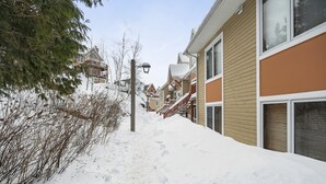 A snow-covered residential street with houses, a wooden fence, and a lamp post. - Hauts-Bois Ski In/Out 2brs w/Pool Access (197-9) (Mont-Tremblant)
