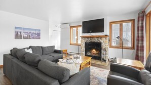 A living room with a stone fireplace, grey sofas, a flat-screen TV, and a large window with blinds. - Hauts-Bois Ski In/Out 2brs w/Pool Access (197-9) (Mont-Tremblant)
