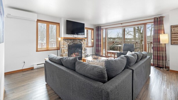 A modern living room with a stone fireplace, a flat-screen TV, and a large window with blinds. - Hauts-Bois Ski In/Out 2brs w/Pool Access (197-9) (Mont-Tremblant)