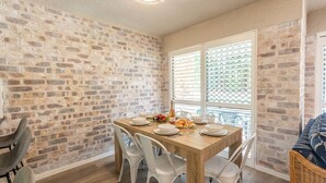 Un coin repas avec un mur en briques, une table en bois dressée pour quatre personnes, des chaises blanches et une fenêtre avec des stores.