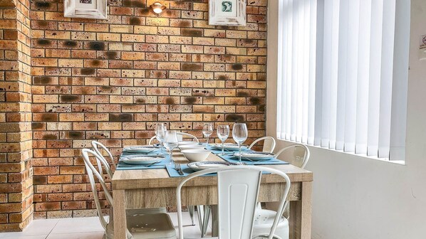 A dining area with a brick wall, a wooden table set with plates and glasses, and white chairs.