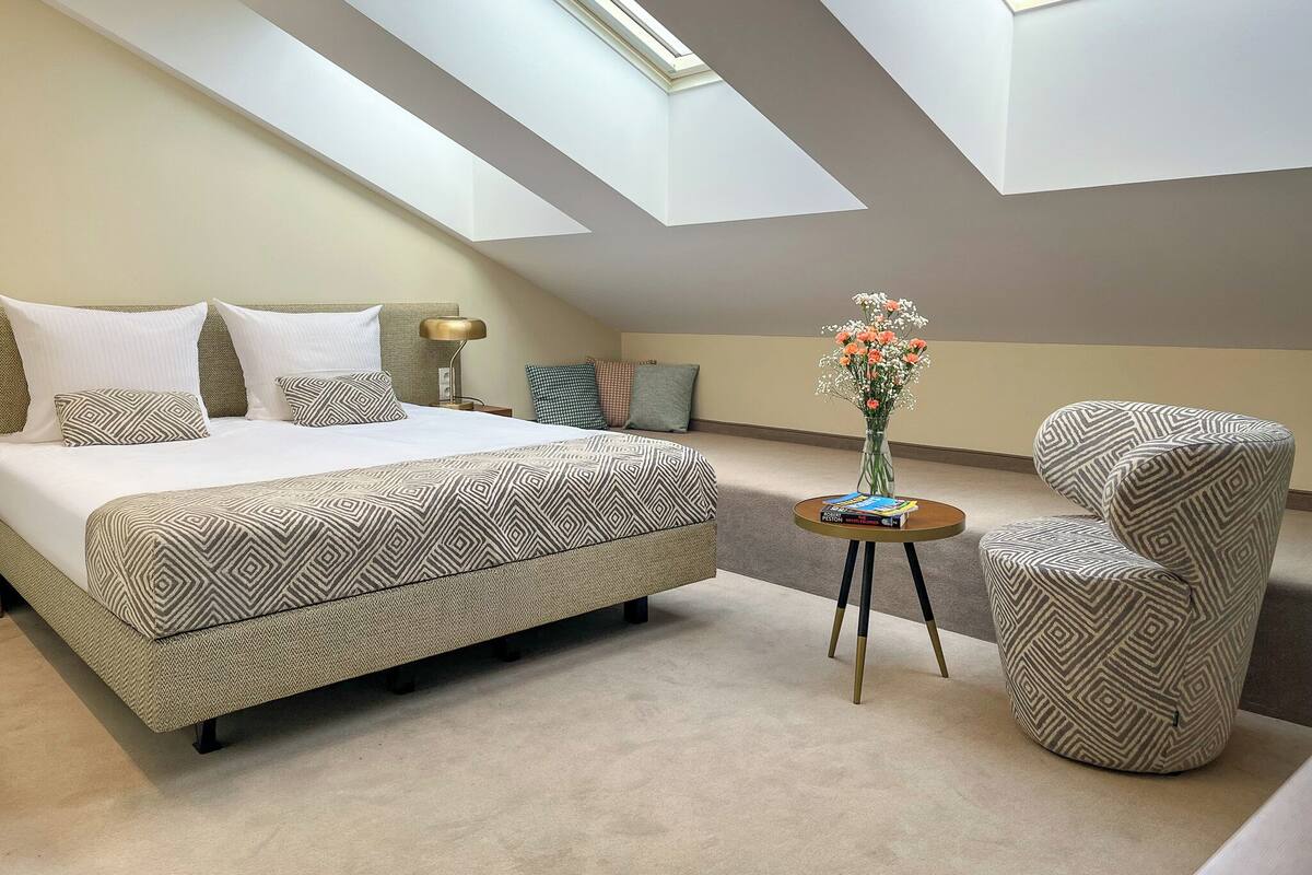 A bedroom with a large bed, a side table with books, a chair, and a skylight.