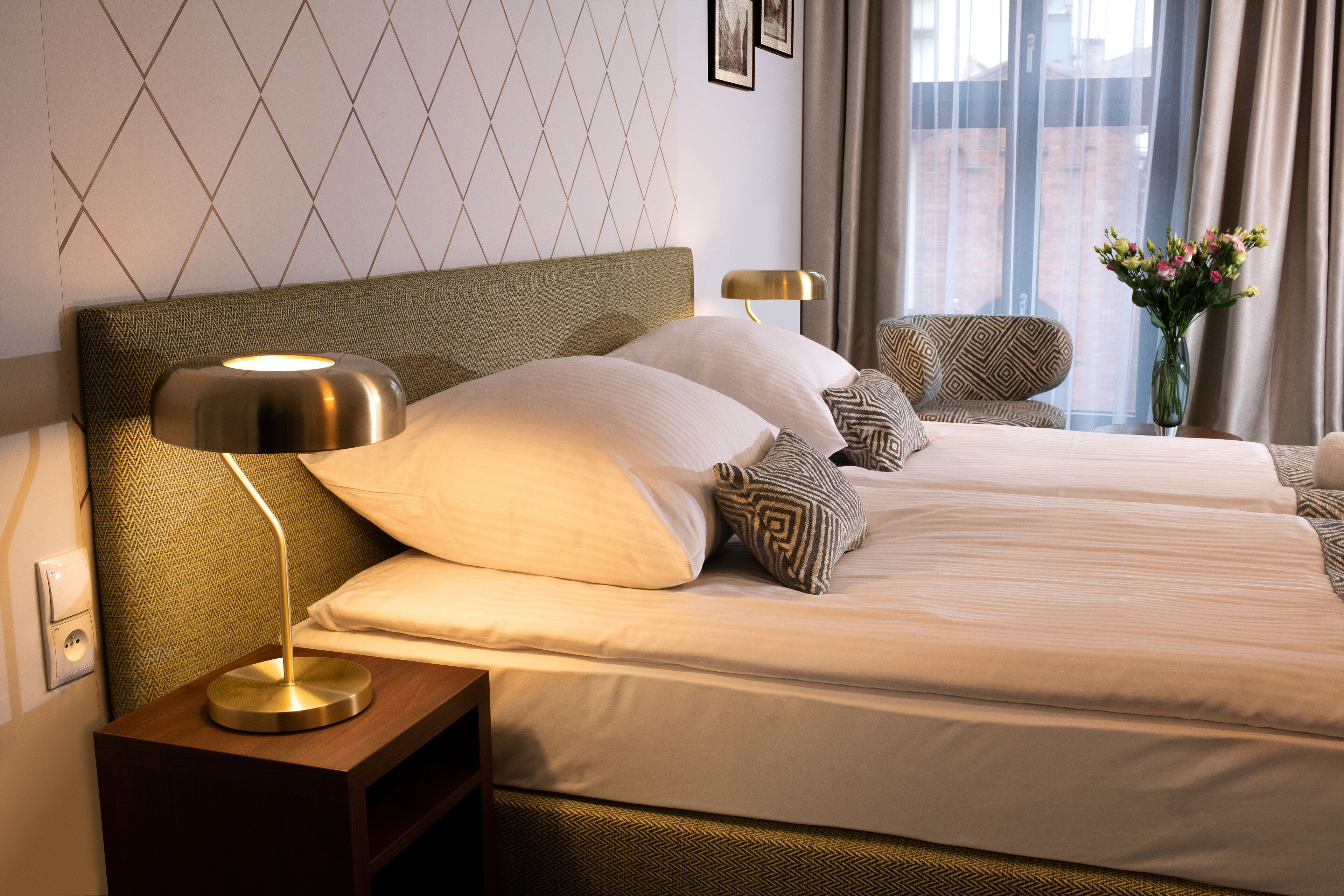 A hotel room with a large bed, a nightstand with a lamp, patterned pillows, and a vase of flowers.