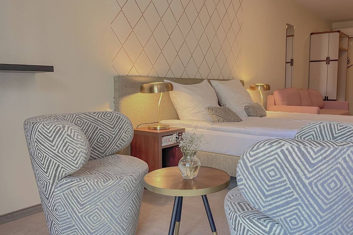 A hotel room with a bed, two patterned armchairs, a small round table, a nightstand, and a wall-mounted lamp.