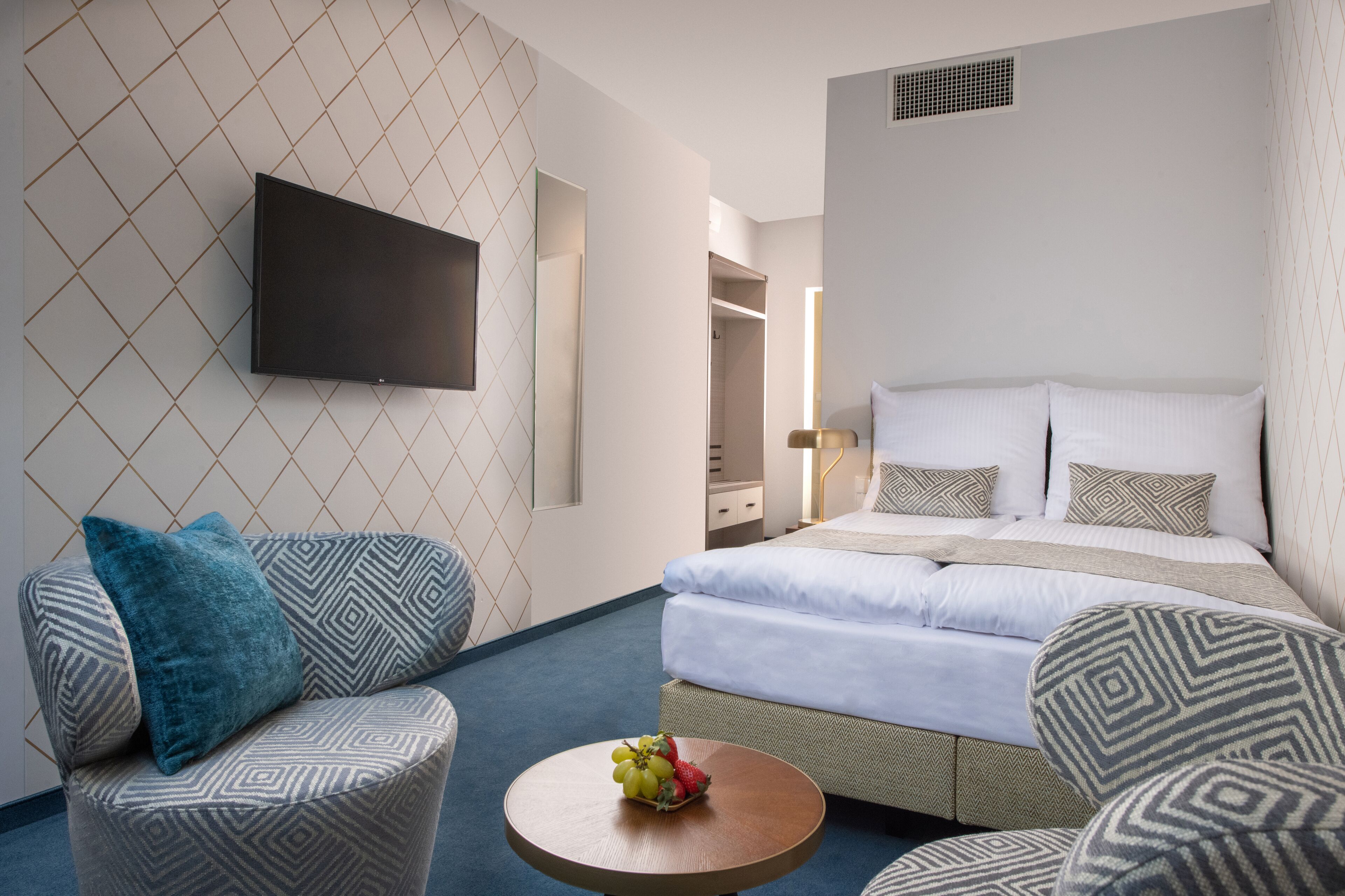 A modern hotel room with a bed, two patterned armchairs, a round coffee table with fruit, and a flat-screen TV on the wall.