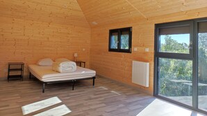 A wooden cabin room with a bed, a small table, a lamp, and a window with a view of greenery.
