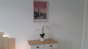 Una habitación con pared blanca, un cartel enmarcado, un mueble blanco pequeño con cajones y una estantería de madera.