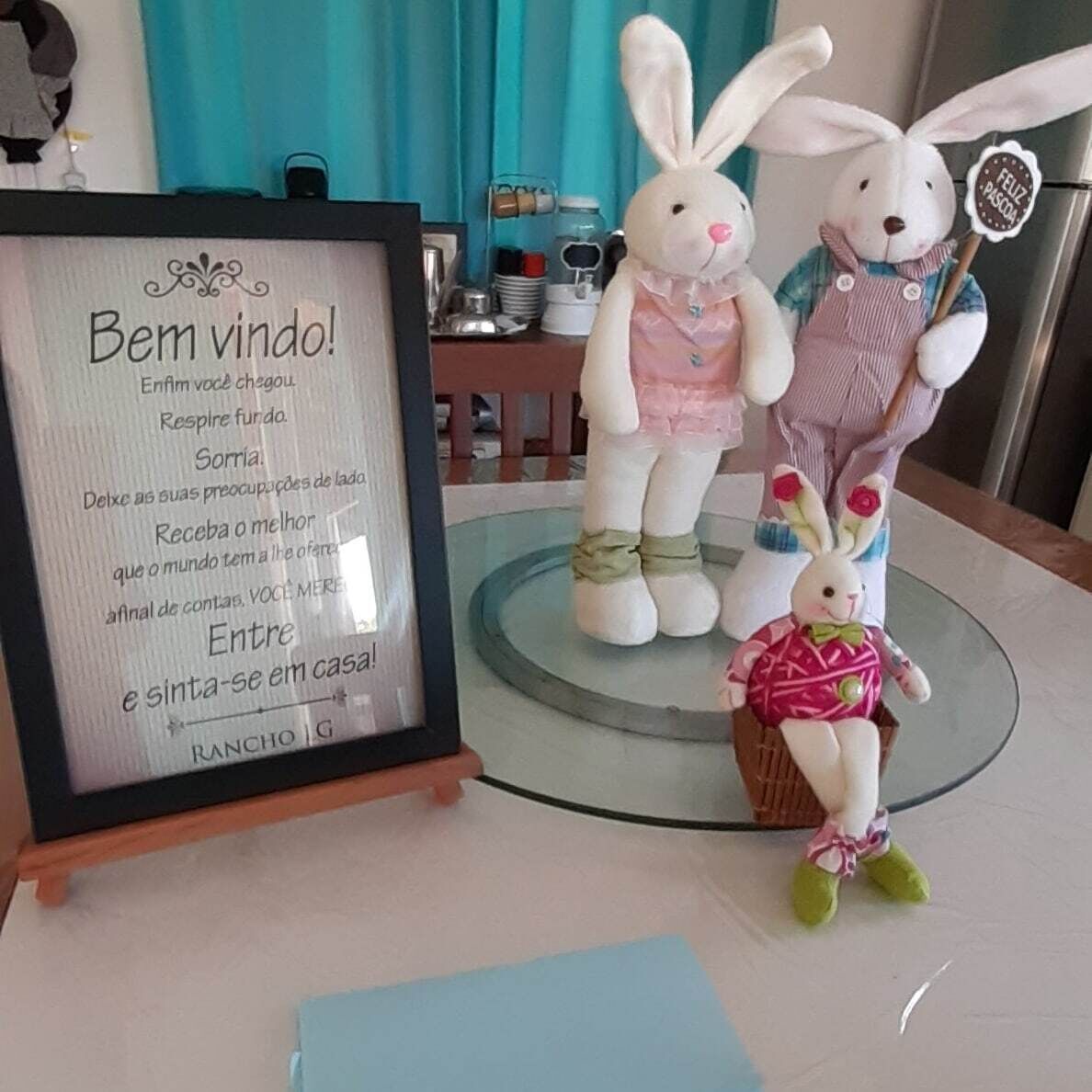 A framed sign with a message in Portuguese, two white plush rabbits, and a small basket with a pink plush toy on a glass table.