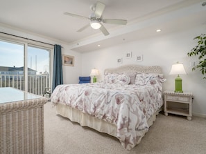 Quarto com cama, ventoinha no teto, mesa de cabeceira com abajur, cadeira de vime e vista para o exterior.