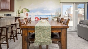 Een eethoek met een houten tafel en stoelen, uitzicht op de zee door een groot raam, en een balkon met een hekwerk.