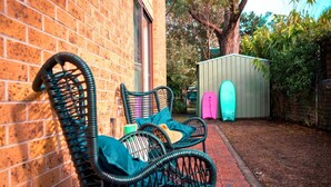 A brick building with a garden area. There is a wicker chair with cushions and a surfboard leaning against the wall. - Gidgets on Gretel - pet-friendly coastal cottage near Shoal Bay (Nelson Bay)