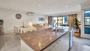 A modern kitchen with a marble countertop, stainless steel sink, and white cabinetry. There is a dining area with a table and chairs, and a living room with a sofa and a large window. - Starfish Sands in Cowes (Cowes)
