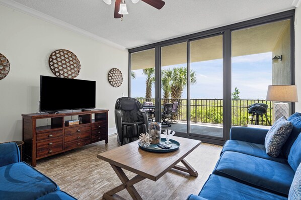 A living room with a TV, wooden furniture, a blue sofa, and a balcony with a view of palm trees.