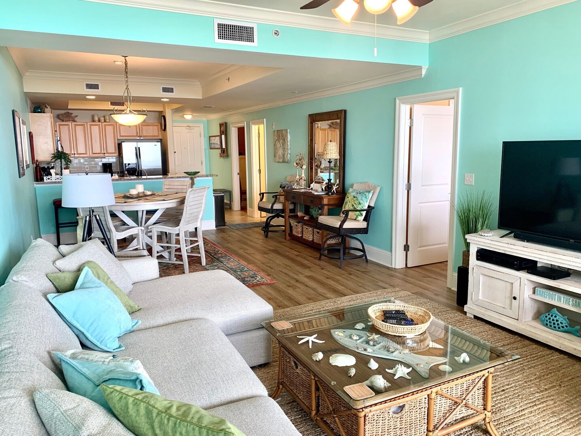 A living room with a sofa, coffee table, and TV stand. There is a kitchen in the background with cabinets and a dining area.