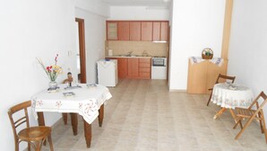 Uma sala de jantar com mesa e cadeiras, uma cozinha ao fundo e uma mesinha com cesta e planta.
