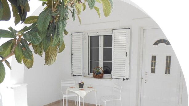 Um quarto branco com chão de azulejos, uma mesa com cadeiras, uma janela com portaria e uma porta com uma pequena janela.