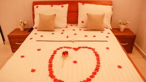 A neatly made bed with rose petals arranged in a heart shape, two pillows, and a vase of flowers on each nightstand. - Residence Appartements Luxueux - Angre - Abidjan (Abidjan)