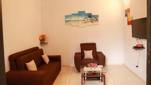 A living room with a brown sofa, a smaller armchair, a glass coffee table, and a wall-mounted TV. - Residence Appartements Luxueux - Angre - Abidjan (Abidjan)