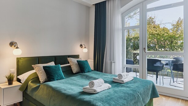 Ein Schlafzimmer mit grüner Bettdecke, zwei Nachttischlampen, einem Nachttisch mit Pflanze und Blick auf eine Außenpatio mit Stühlen.