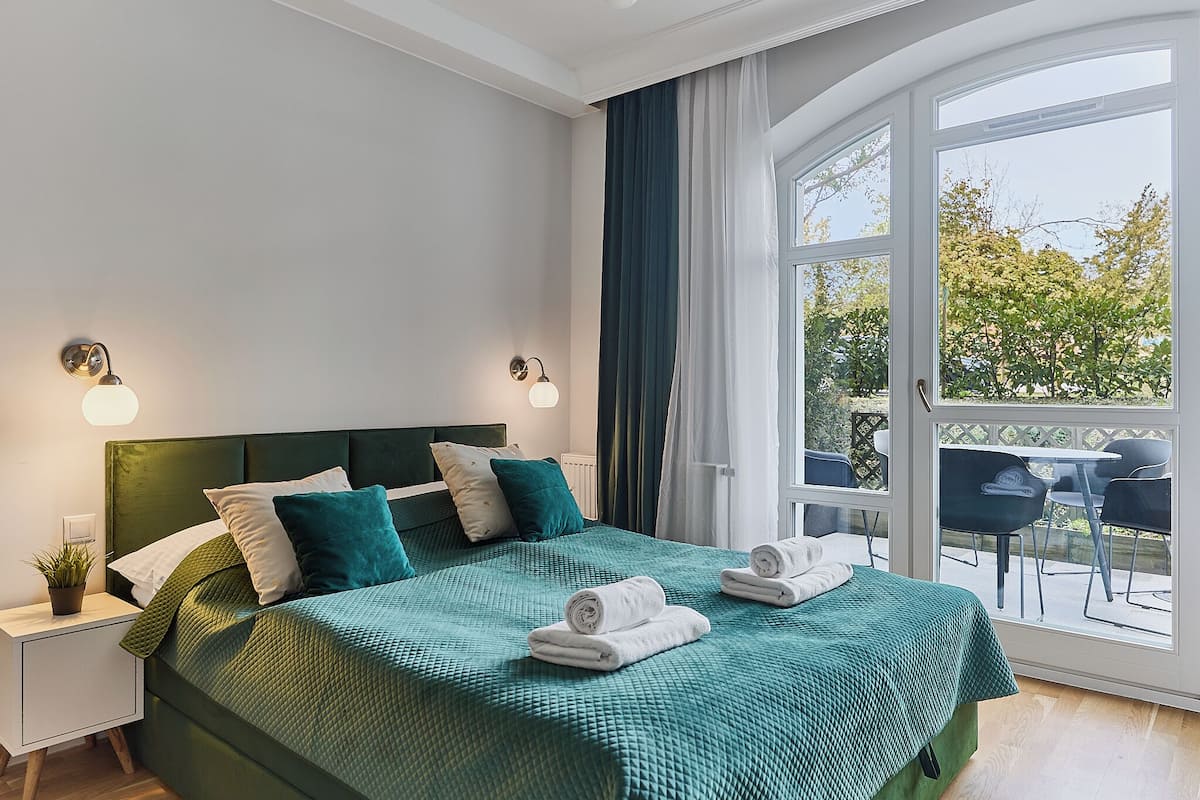 A bedroom with a green bedspread, two bedside lamps, a nightstand with a plant, and a view of an outdoor patio with chairs.
