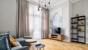 Ein modernes Wohnzimmer mit einem grauen Sofa, zwei kleinen runden Tischen, einem Fernseher auf einem Holzständer und einer schwarzen Regaleinheit mit Dekorationselementen.