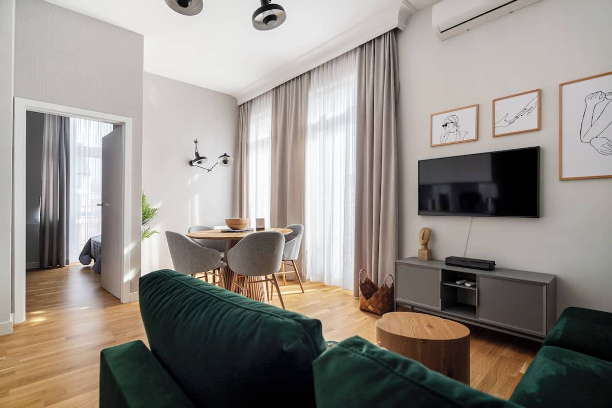 A modern living room with a green sofa, wooden flooring, a dining area with chairs, a TV, and a grey cabinet.