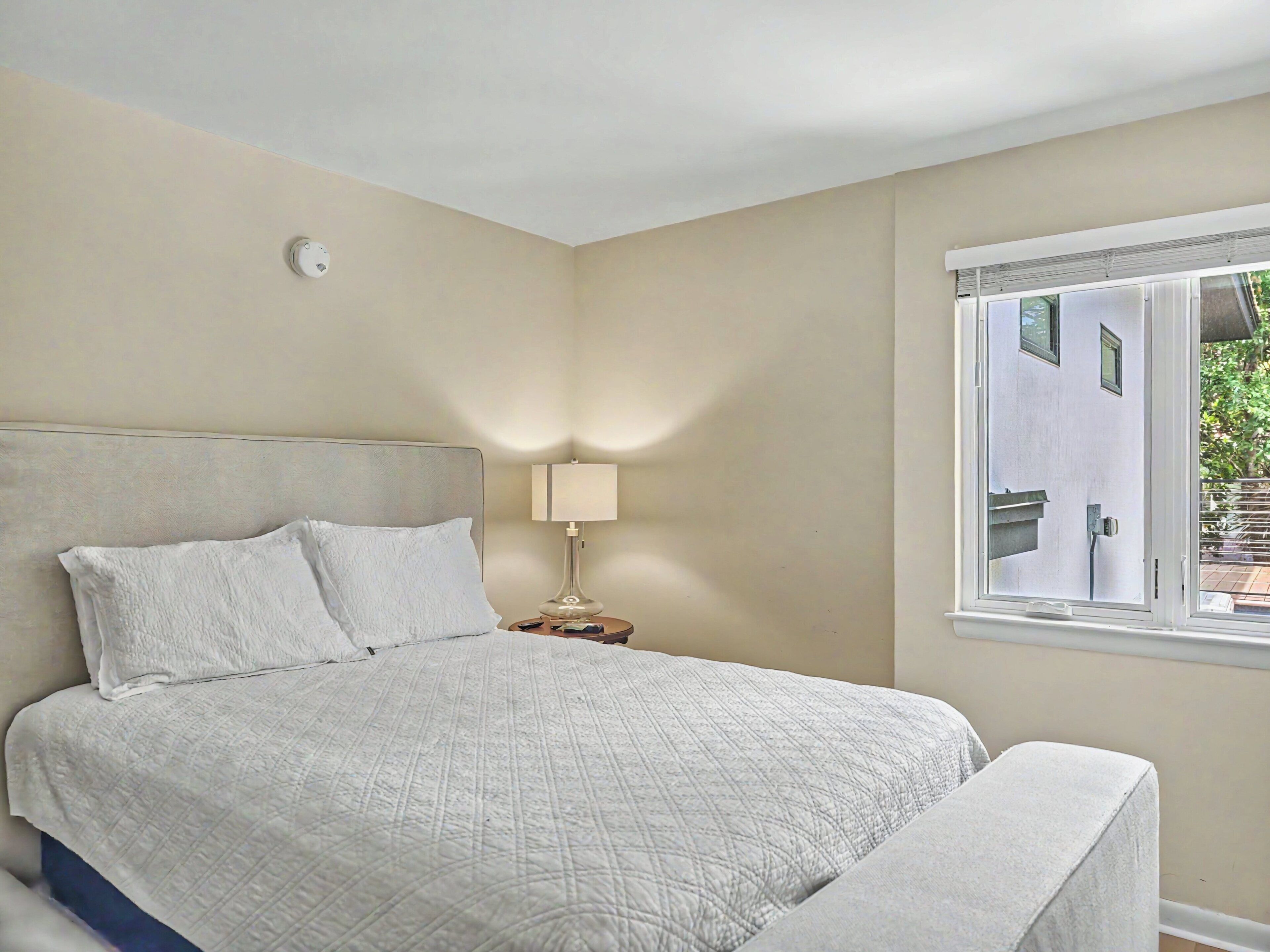 A bedroom with a bed, a bedside lamp, a window, and a wall-mounted light fixture.