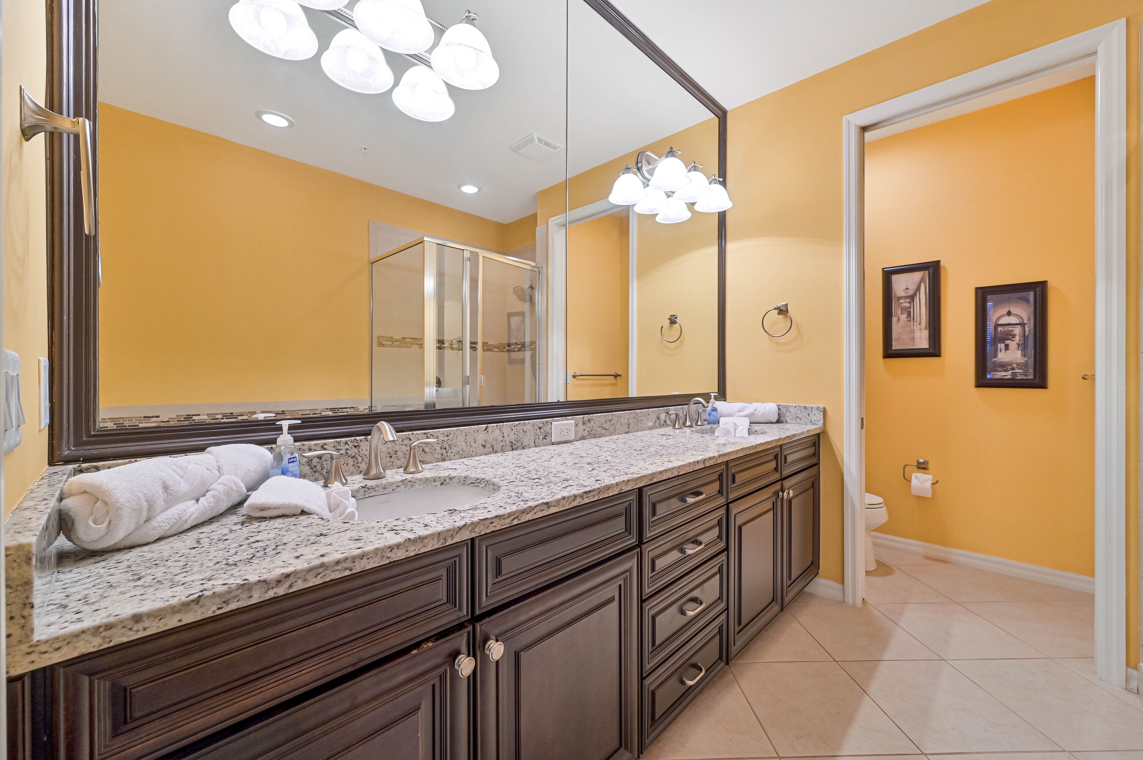 A bathroom with a large mirror, dark wooden cabinets, a marble countertop, and a toilet.
