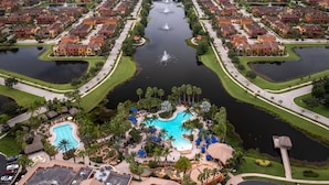 Une vue aérienne d’un quartier résidentiel, avec un élément aquatique central bordé de maisons, d’une piscine et d’un petit espace vert aménagé avec des chaises longues.