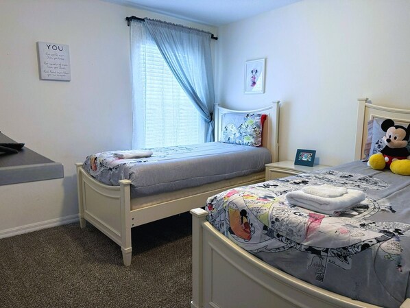 A bedroom with two single beds, a window with curtains, a Mickey Mouse plush toy, and a motivational quote on the wall.